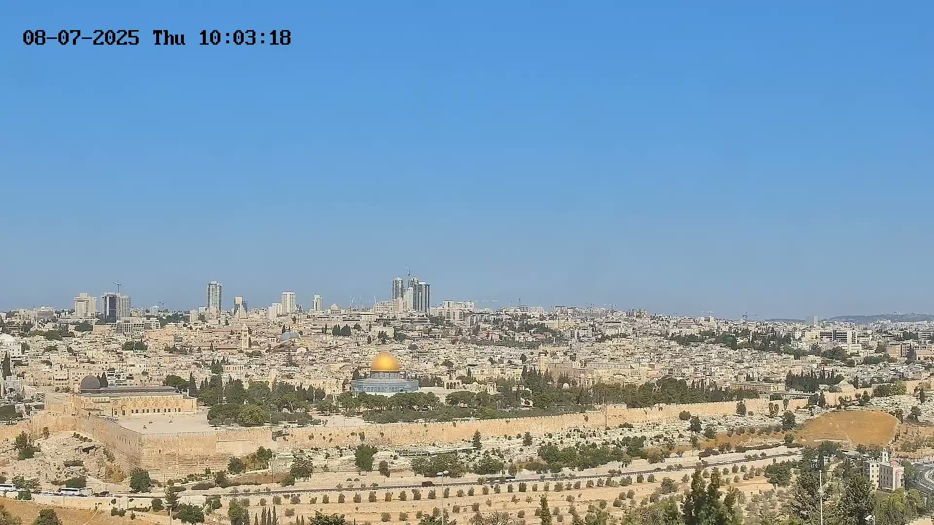 A wide, long shot shows a sprawling city under a clear blue sky, with a prominent gold dome structure at its center.