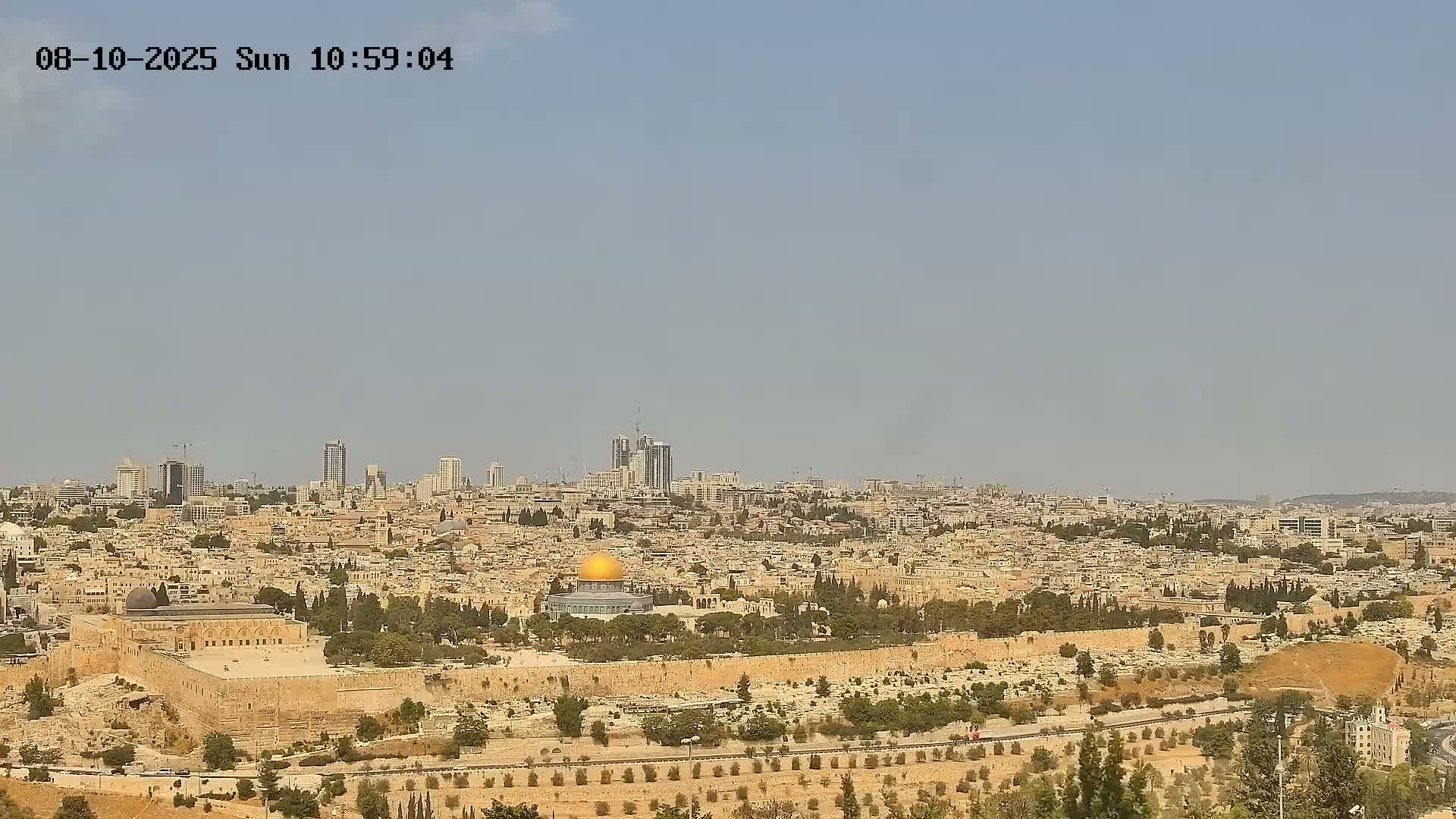 A distant view of Jerusalem on a clear, sunny day shows the Dome of the Rock within the city walls, surrounded by buildings extending to a skyline of taller structures.
