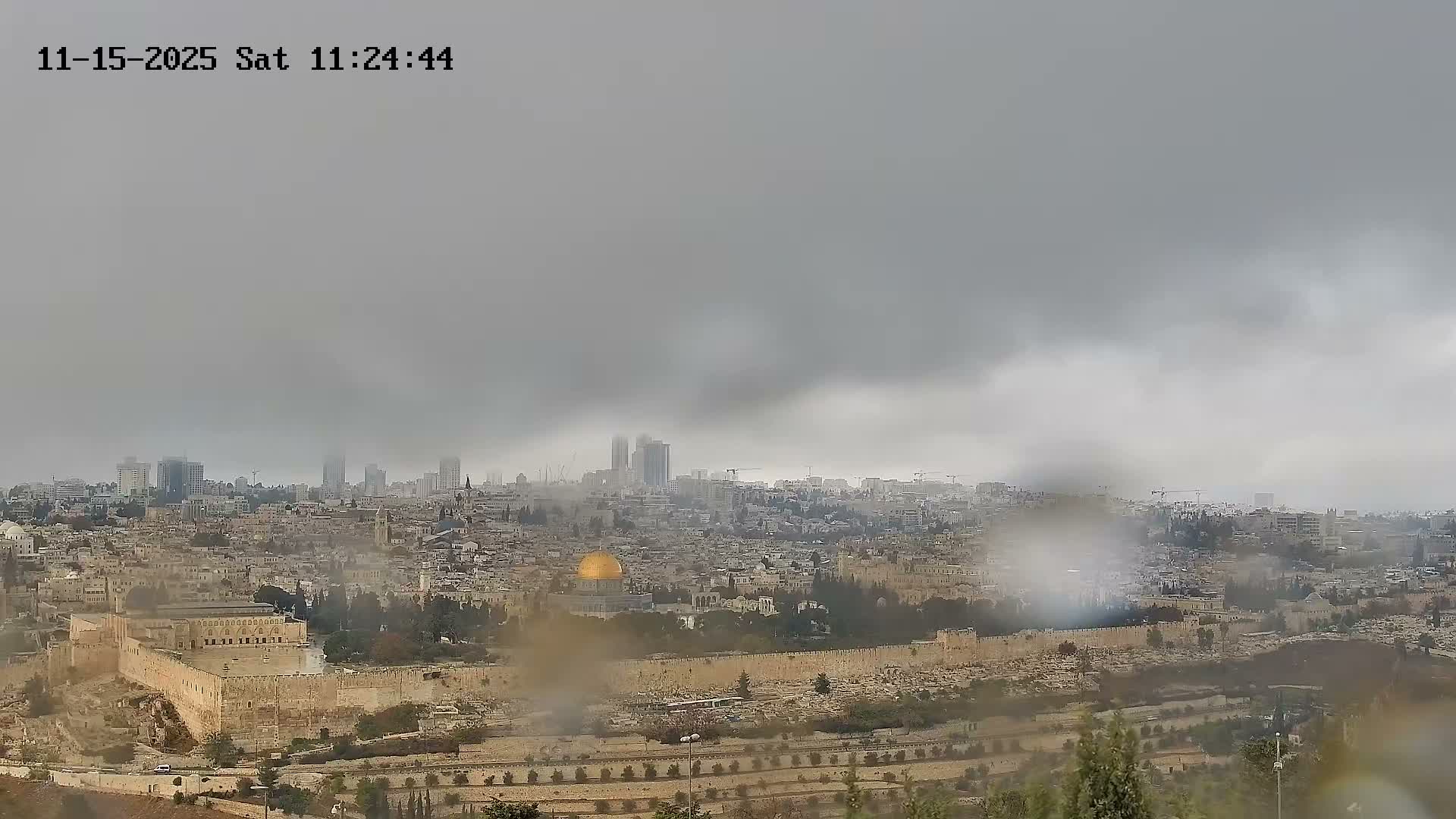 A panoramic view of Jerusalem's Old City, featuring the prominent golden Dome of the Rock and Al-Aqsa Mosque, is seen under a heavily overcast and hazy sky, with visible rain or smudges on the camera lens obscuring parts of the cityscape.