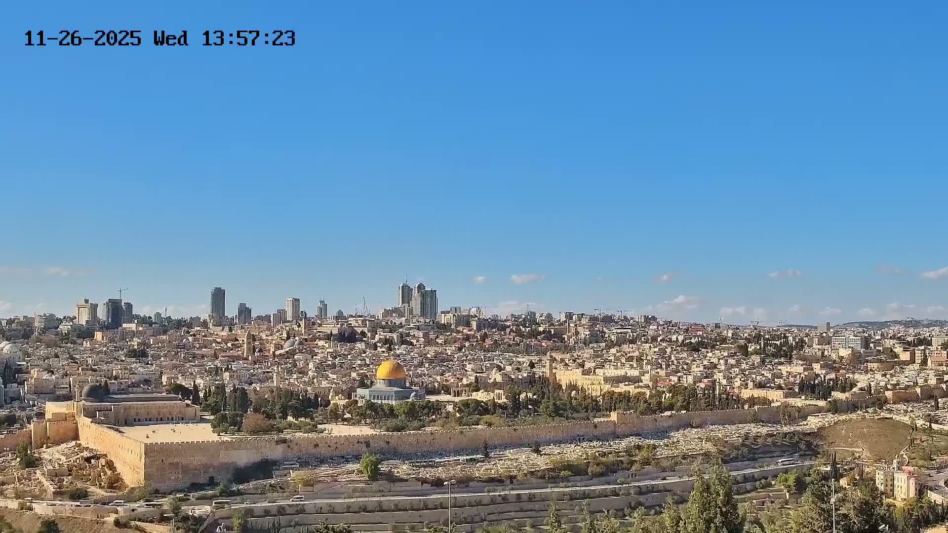 A sprawling panoramic view of a densely packed historic city, featuring the golden dome of the Dome of the Rock and the Al-Aqsa Mosque complex prominently in the midground, with a large cemetery on a hillside to the right and modern high-rises in the hazy distance, all under a clear blue, sunny sky.