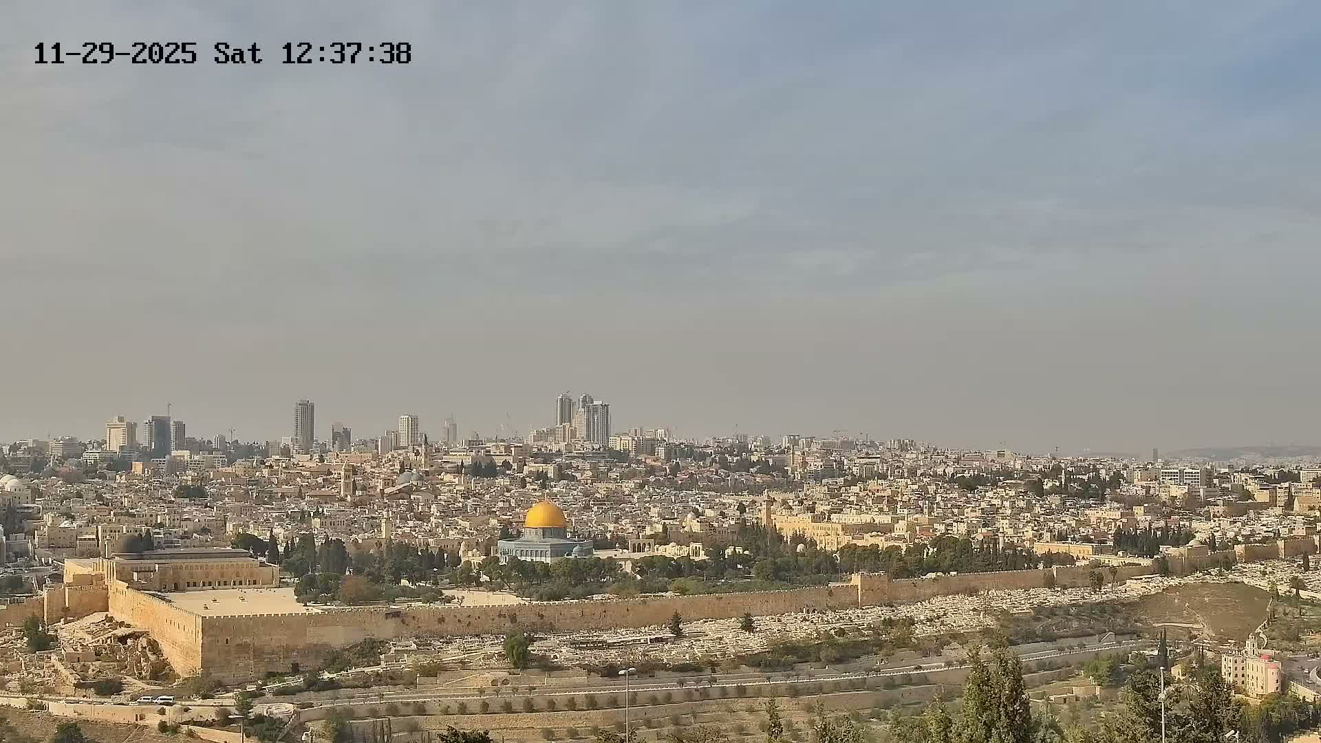 A wide view of Jerusalem reveals the historic Old City walls, the golden Dome of the Rock, and the Al-Aqsa Mosque amidst a sprawling urban landscape of both ancient and modern buildings, all under a hazy, partly cloudy sky.