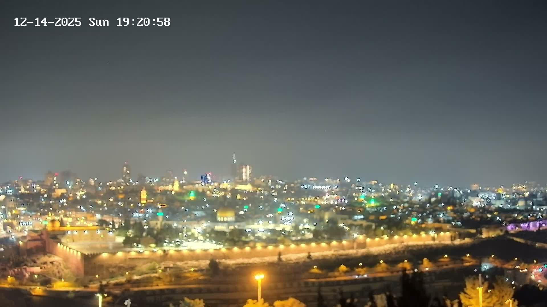 An expansive panoramic view captures a historic city under a partly cloudy, bright sky, featuring the prominent golden Dome of the Rock and Al-Aqsa Mosque nestled within ancient walls, surrounded by a densely built urban landscape extending into the distance.
