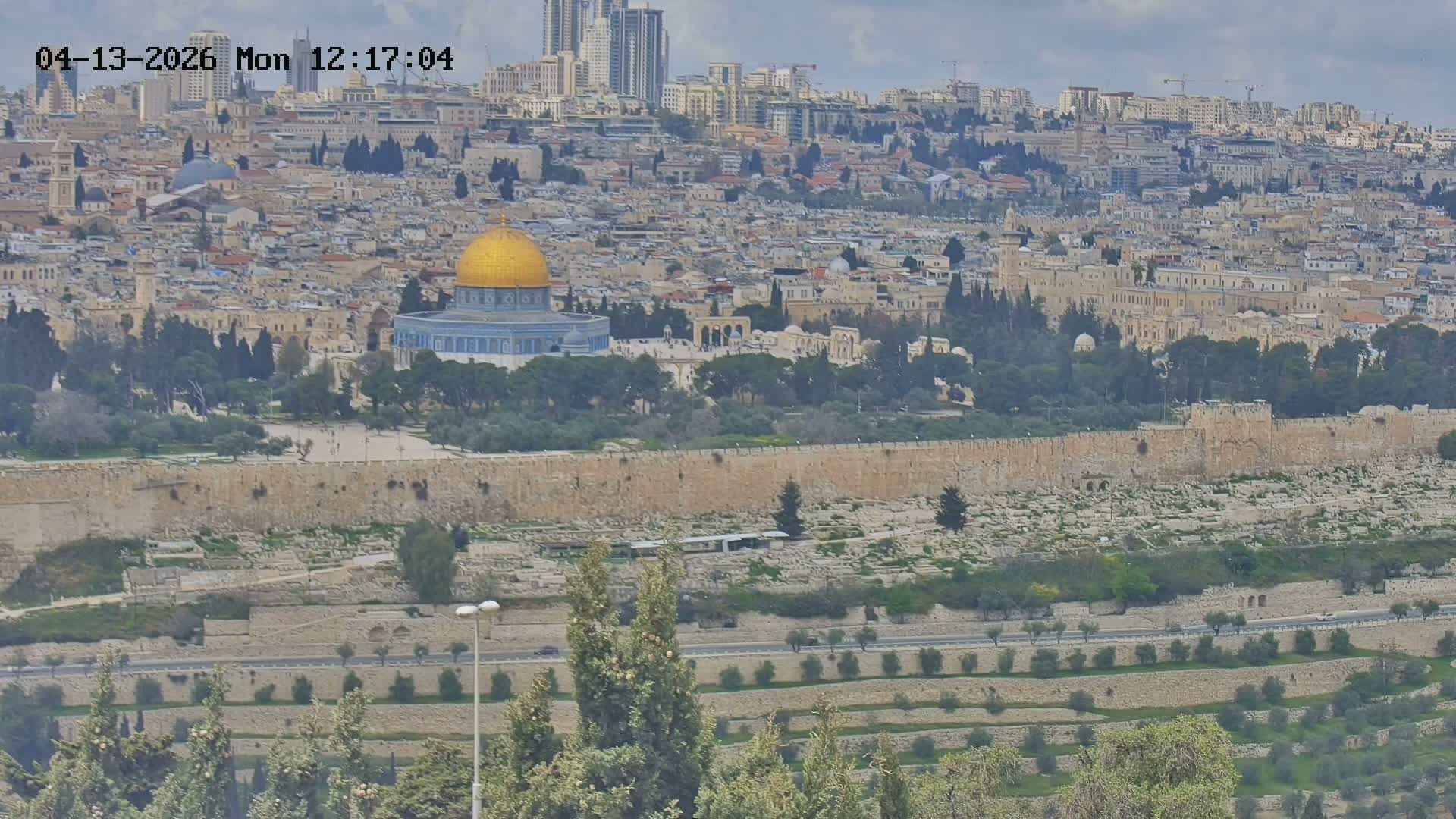 An expansive view of a sunny cityscape reveals the historic walled city of Jerusalem, including the golden-domed Dome of the Rock and Al-Aqsa Mosque, surrounded by numerous buildings extending to modern high-rises under a clear blue sky.