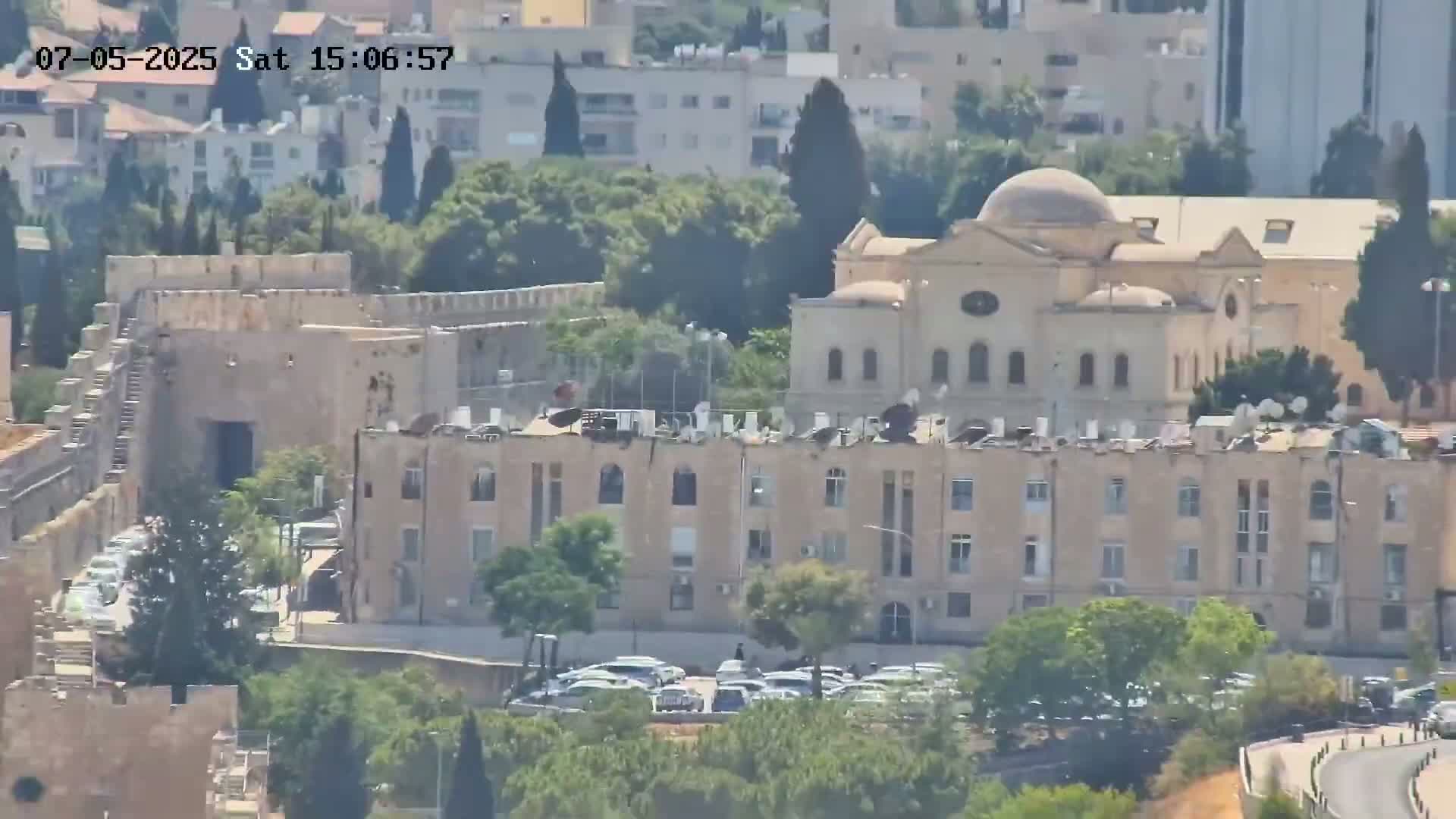 A high-angle, long shot shows a stone wall, a multi-story building, and a domed building on a sunny day.