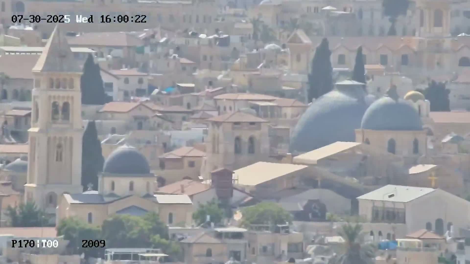 A high-angle, long shot on a sunny day shows a sprawling city with ancient stone walls and structures, including a mosque with a dome, situated next to a road winding through the landscape.