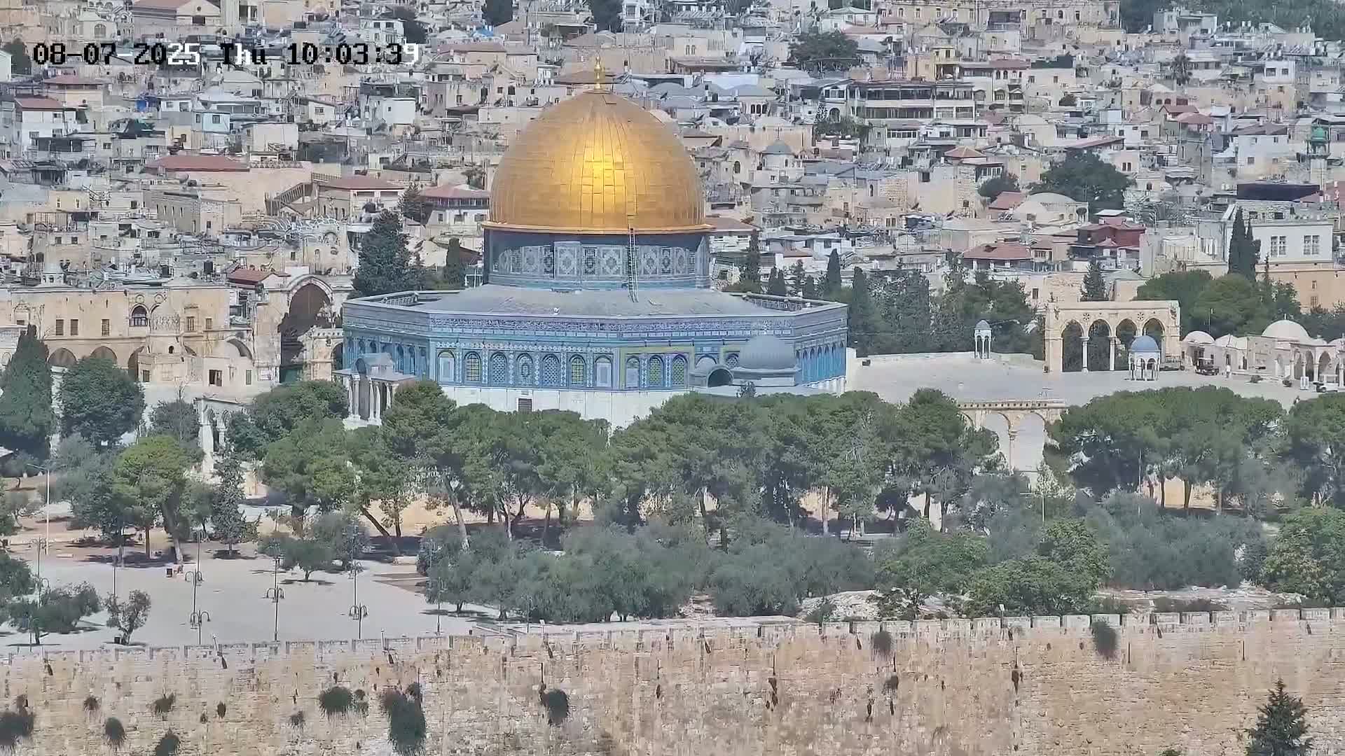 A gold-domed building with blue and white tilework is seen from a distance, surrounded by trees, a courtyard, and a stone wall, under a sunny sky.