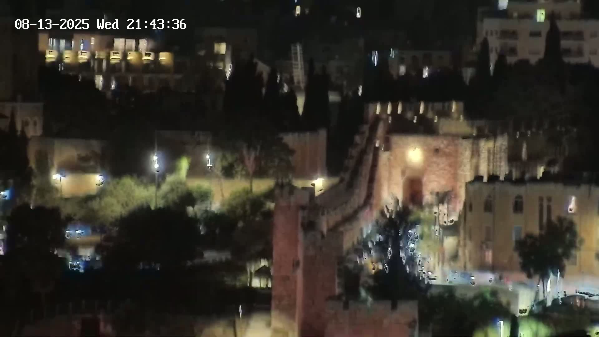 A nighttime, long shot of a stone structure, possibly a castle or fortress, illuminated by artificial lights, with some surrounding vegetation and buildings visible in the background.