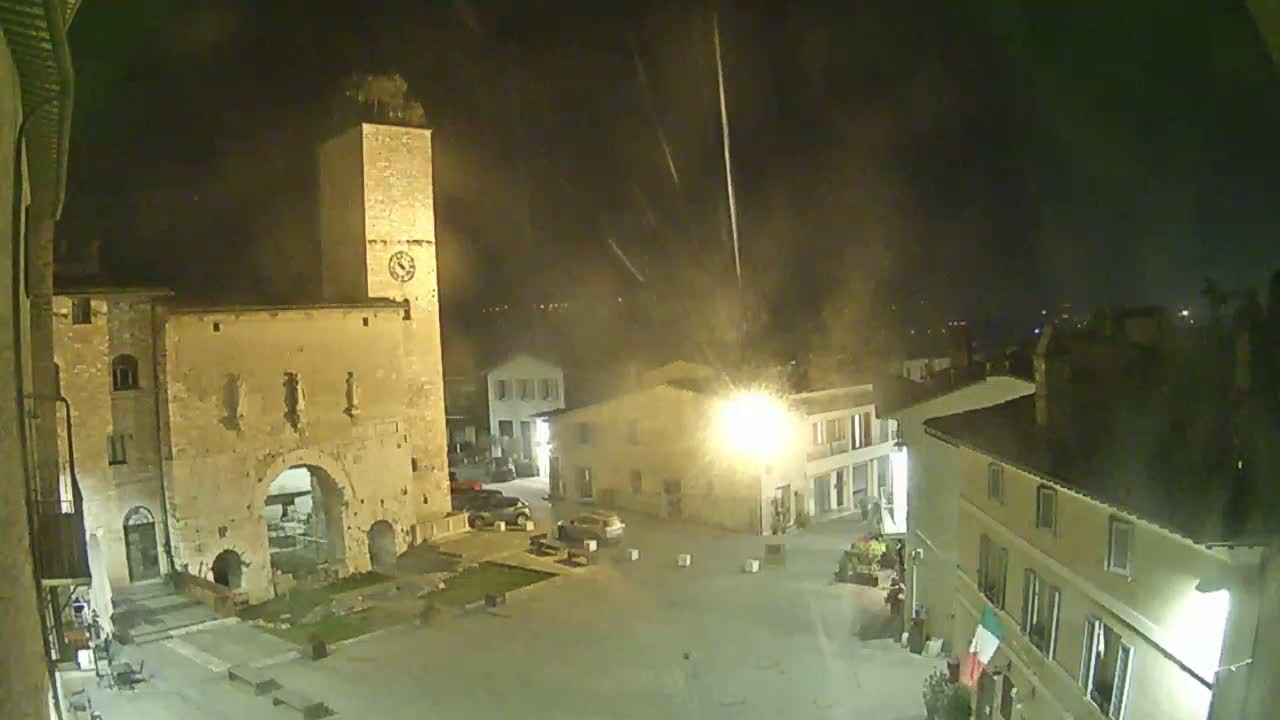 A nighttime view of a town square with ancient stone buildings, including a tall clock tower, under a slightly hazy sky with streaks of light possibly from rain or fireworks.