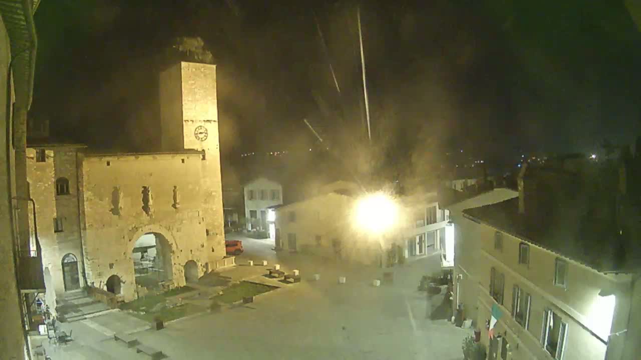 A nighttime, high-angle view shows a town square with old stone buildings, a clock tower, and a few cars, under a hazy sky with streaks of light possibly from fireworks.