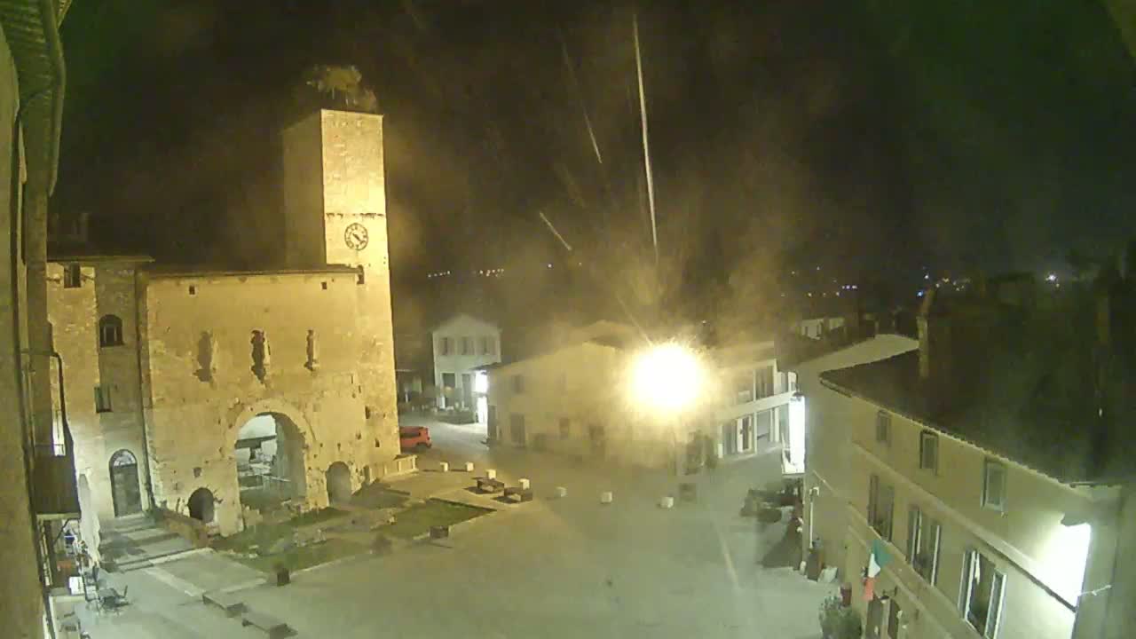 A nighttime view of a town square with an old stone tower and buildings, under conditions of light rain or snow.