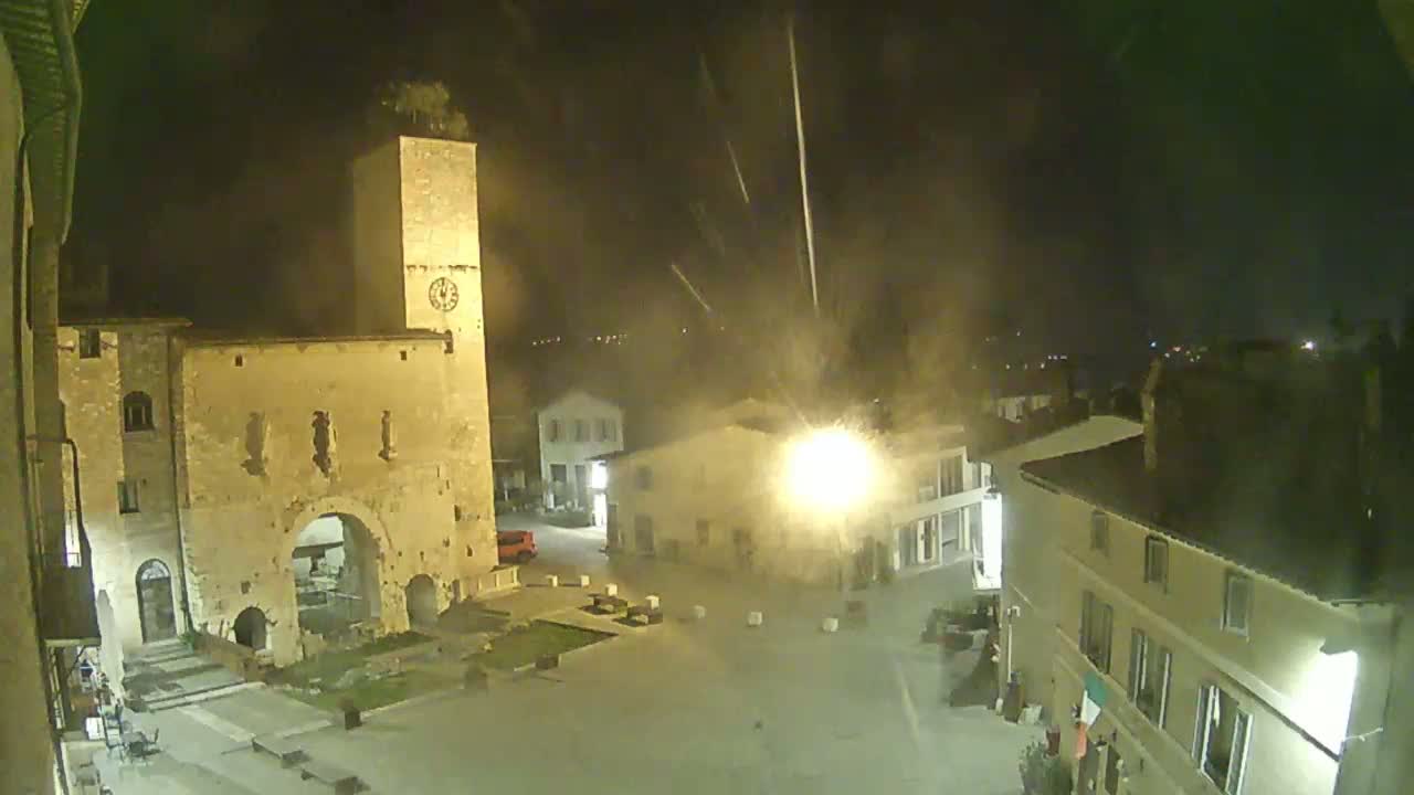 A nighttime view of a town square with stone buildings, a clock tower, and light rain or mist falling.