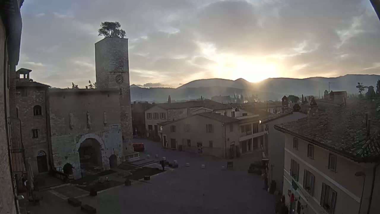 A sunlit town square in a hilly region is surrounded by ancient-looking stone buildings at sunrise under a partly cloudy sky.