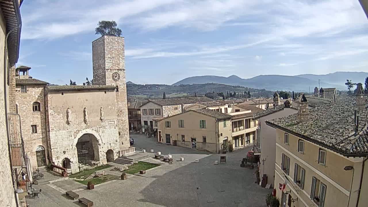 A town square in a small Italian town, under a partly cloudy sky, is surrounded by stone buildings, including a tall tower with a tree on top, and overlooks a hilly landscape in the distance.