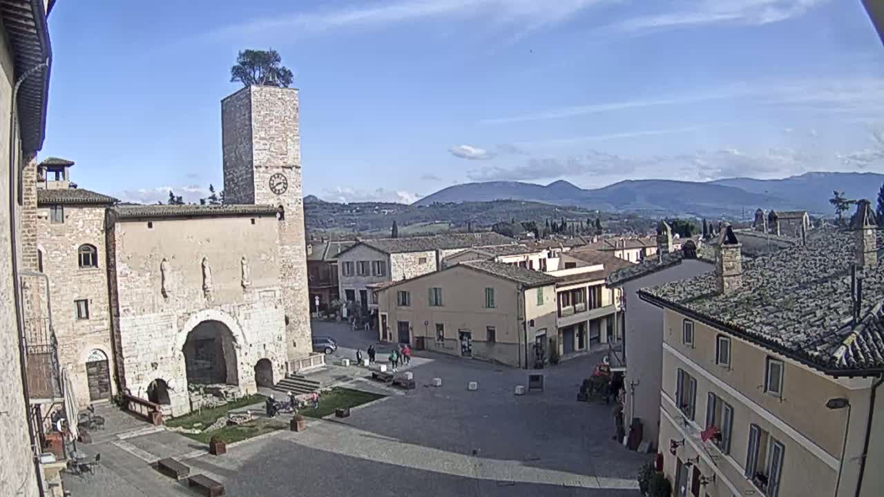 A town square in a quaint European village, under a mostly sunny sky, is surrounded by stone buildings, including a tall clock tower with a tree on top, and overlooks a range of distant hills.