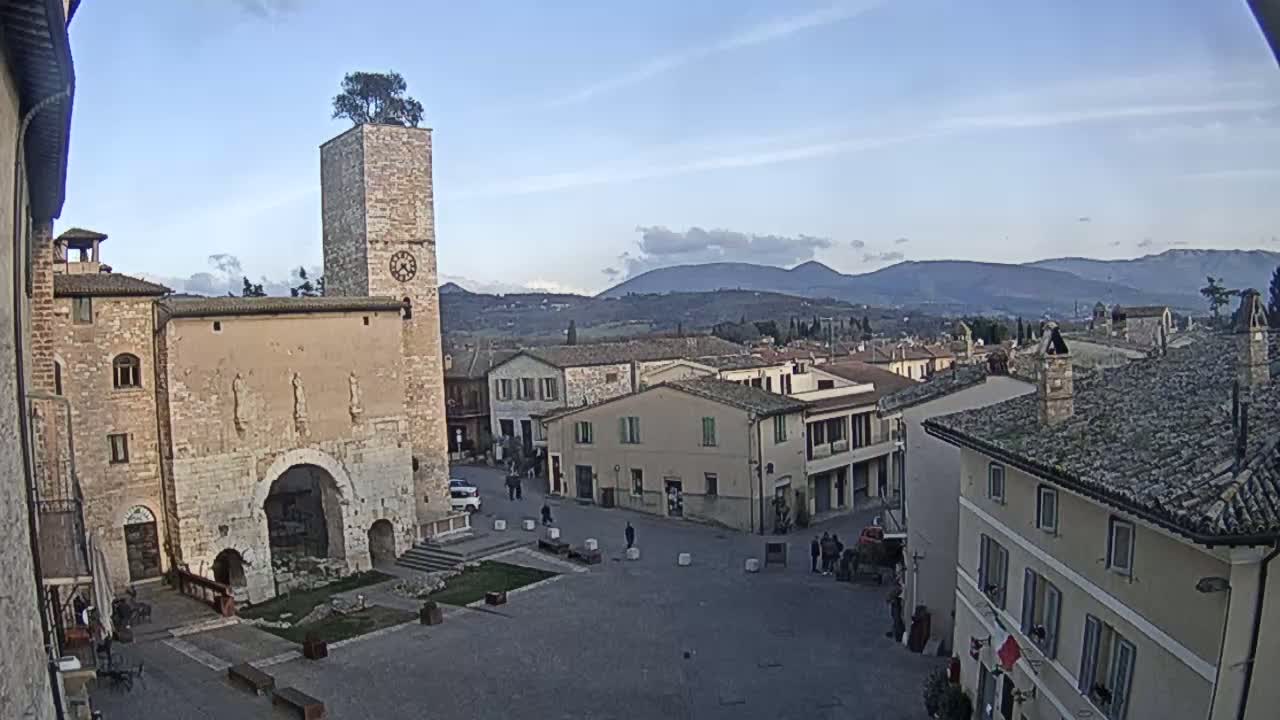 A town square in a historic Italian town, under a mostly clear sky with distant mountains visible in the background.