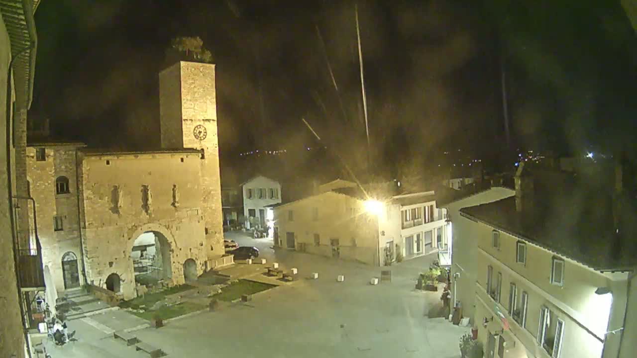 A nighttime view of a town square with ancient stone buildings, including a tall clock tower, under a slightly hazy sky with streaks of light possibly from rain or fireworks.