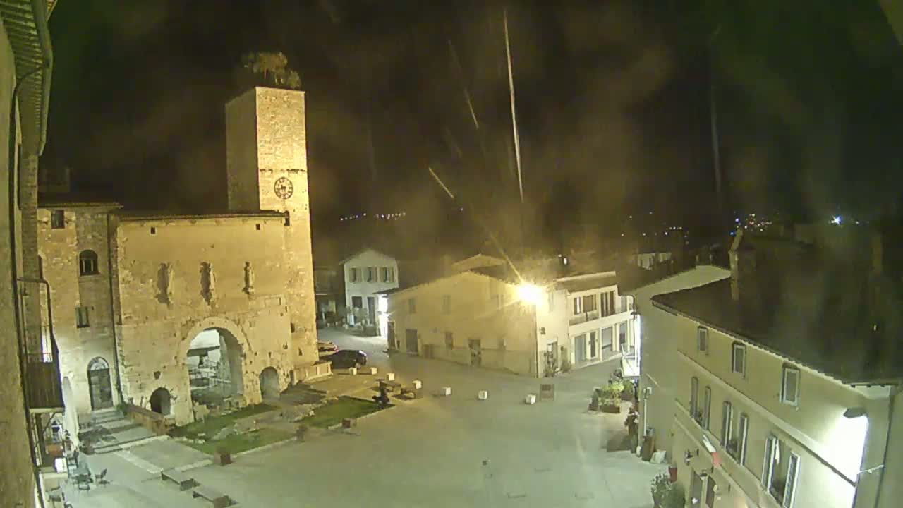An empty town square at night, illuminated by streetlights, is surrounded by old stone buildings, including a tall tower with a clock, under a slightly hazy sky.