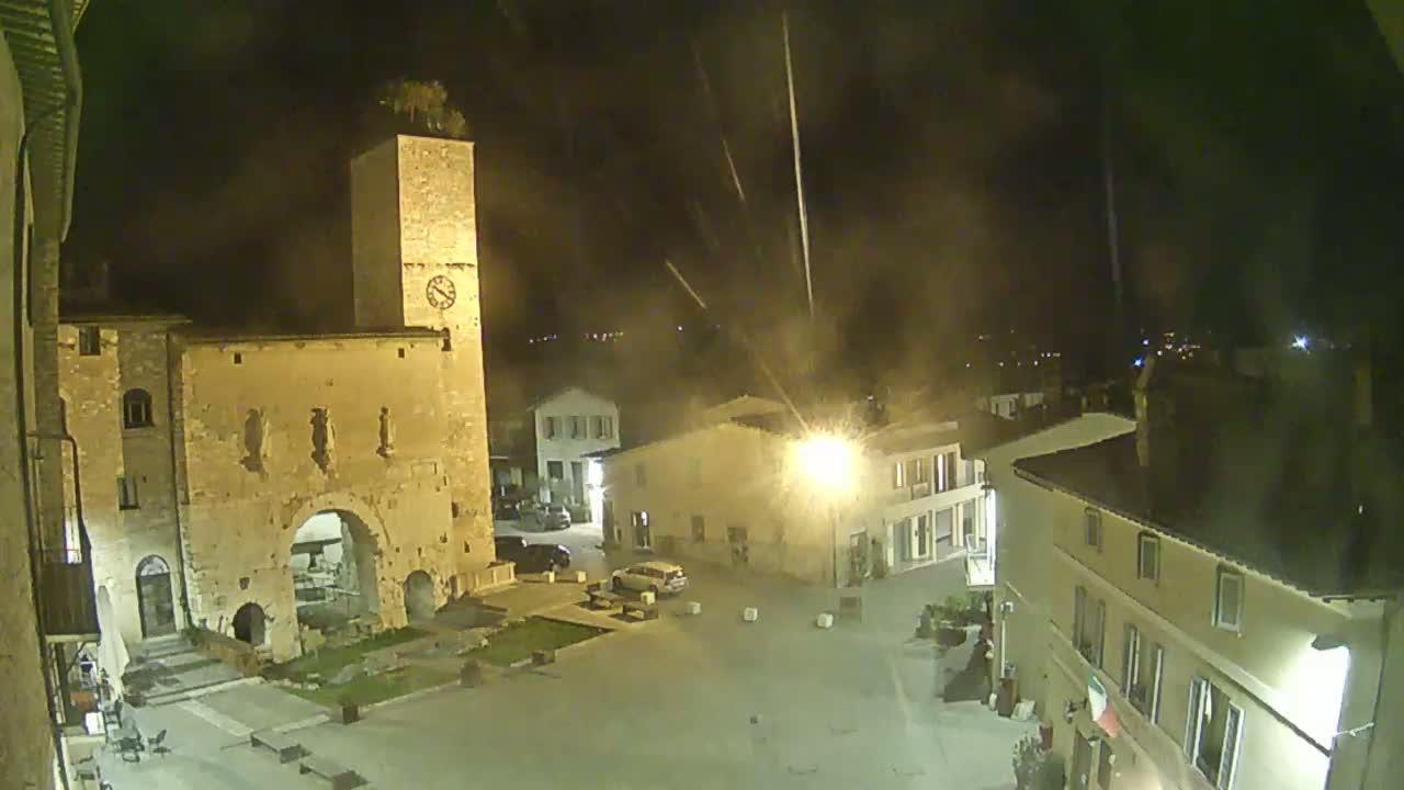 A nighttime view of a town square, featuring ancient stone buildings, a clock tower, and a few cars parked, under a hazy, possibly smoky sky.