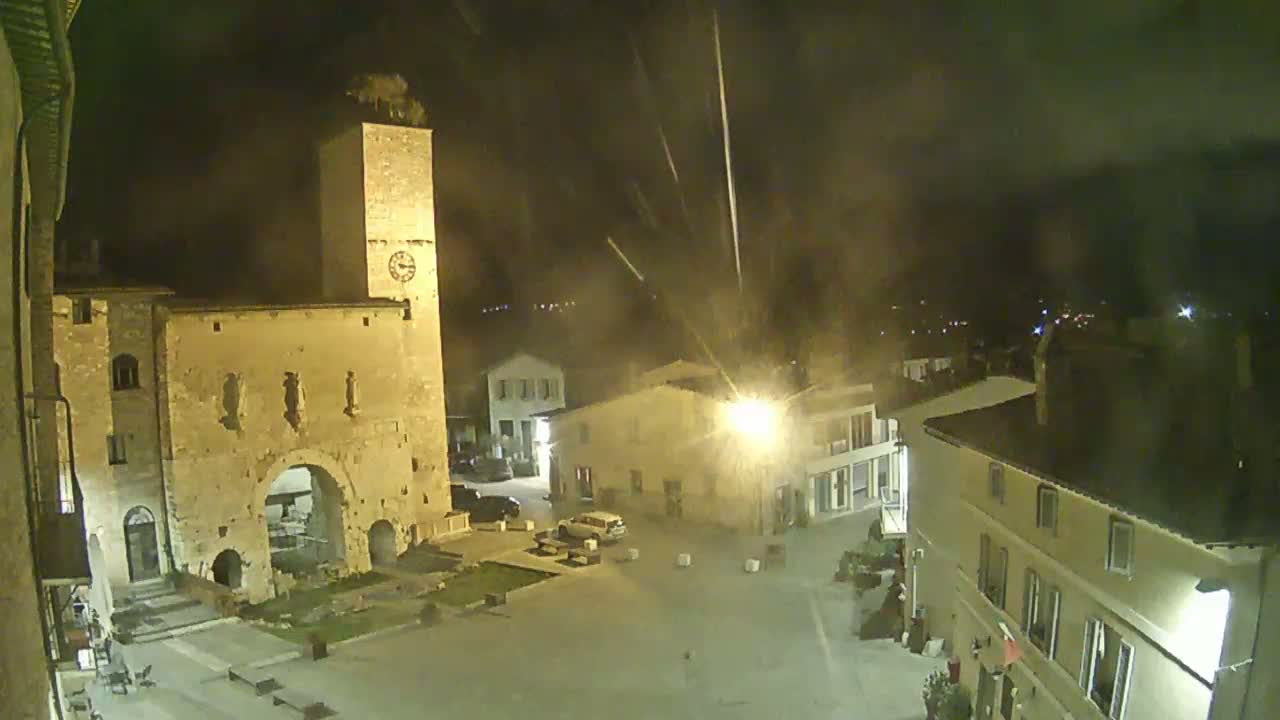 A nighttime view of a town square with stone buildings, a tall clock tower, and faint streaks of light in the sky.