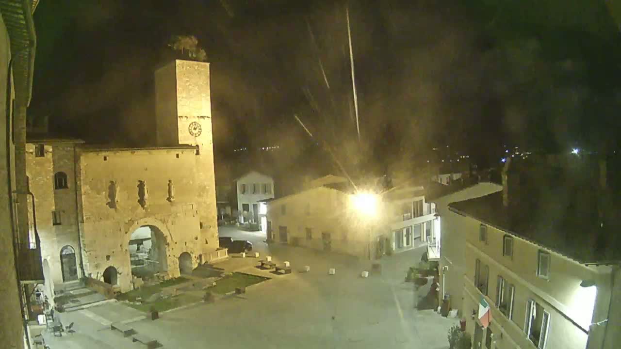 A nighttime view of a town square with old stone buildings, including a clock tower, under a light rain or snow.