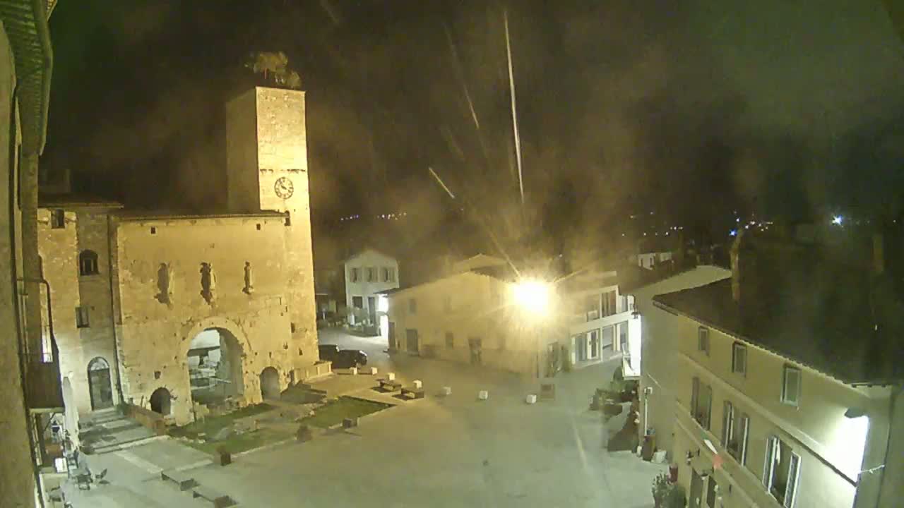 A nighttime view of a town square with a tall stone tower, buildings, and faint streaks of light possibly from rain or snow in the dark sky.