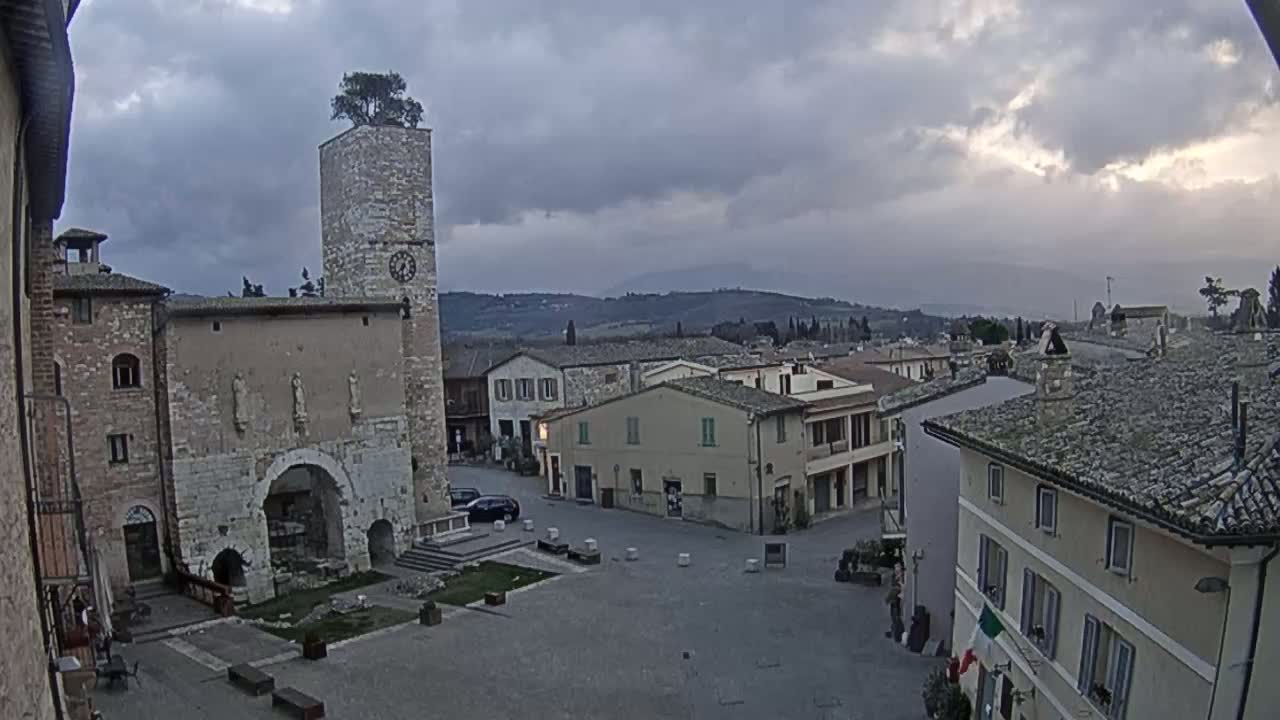 A town square in a stone village under an overcast sky, centered on a tall clock tower with a tree on top, overlooks rolling hills in the distance.