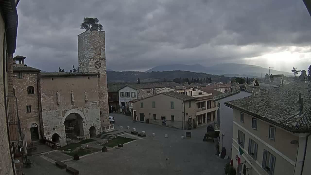 A town square with stone buildings, a clock tower topped with a tree, and distant mountains under a cloudy sky.