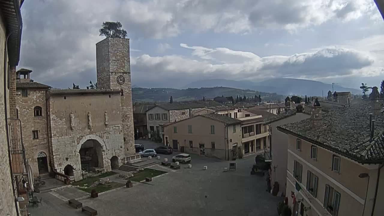 A town square in a small Italian town, under a partly cloudy sky, is surrounded by aged stone buildings, one with a tall clock tower topped with a small tree.