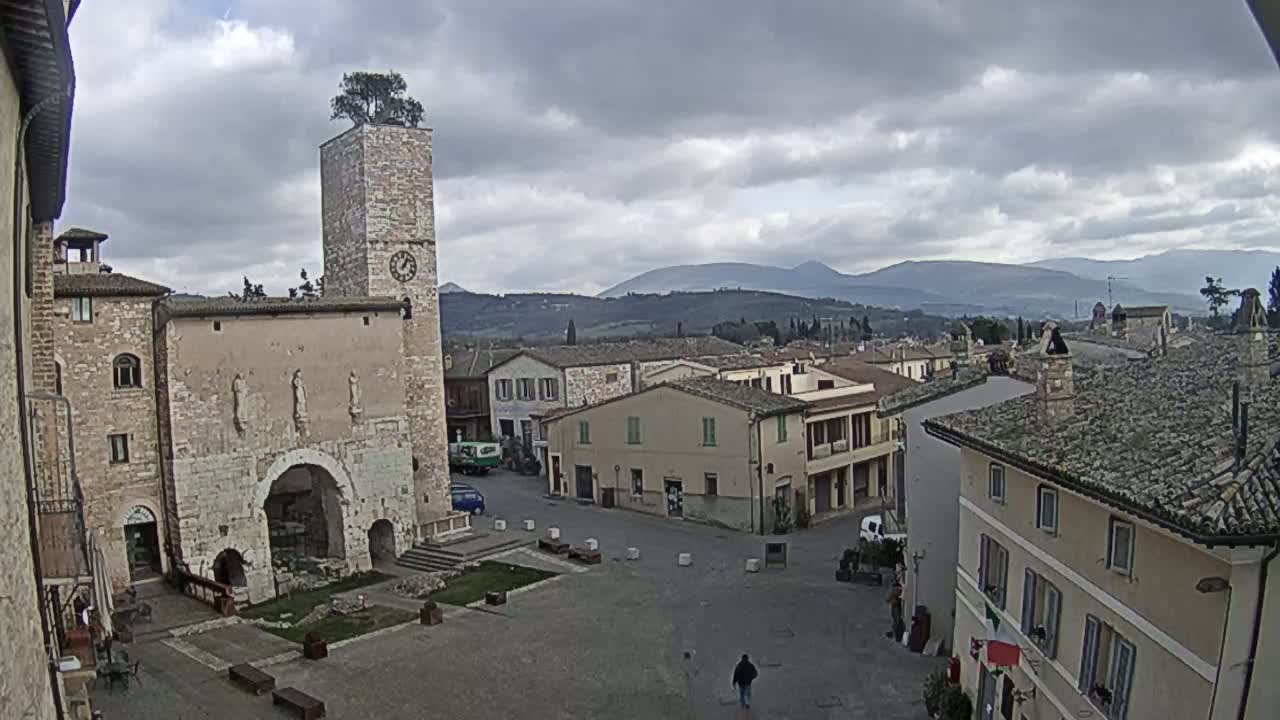A town square with stone buildings, a clock tower topped with a tree, and distant mountains under a cloudy sky.