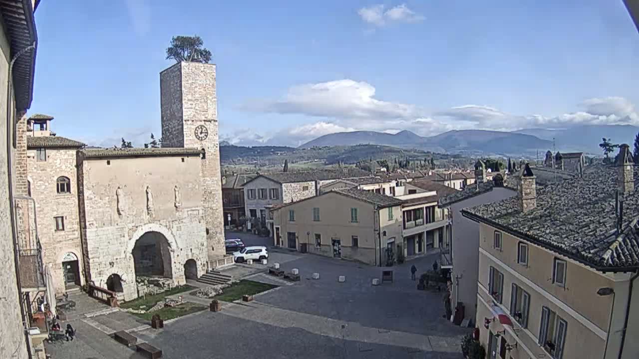 A mostly sunny day reveals a town square in a European-style village, centered on an old stone building with a clock tower topped with a small tree, with distant mountains visible under a partly cloudy sky.