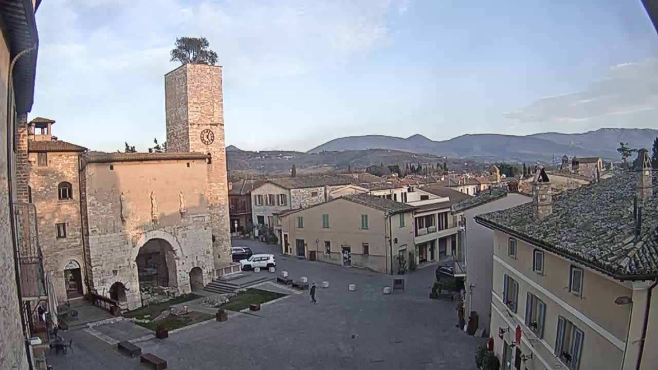 A town square in a stone village, under a mostly clear sky, is surrounded by buildings including a tall tower with a tree on top, and overlooks distant mountains.