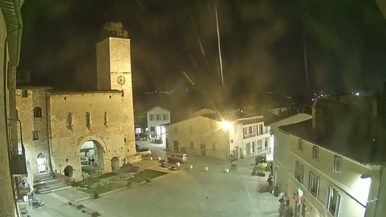 A nighttime view of a town square with an ancient stone archway and clock tower, under a slightly hazy sky with streaks of light possibly from fireworks or meteors.