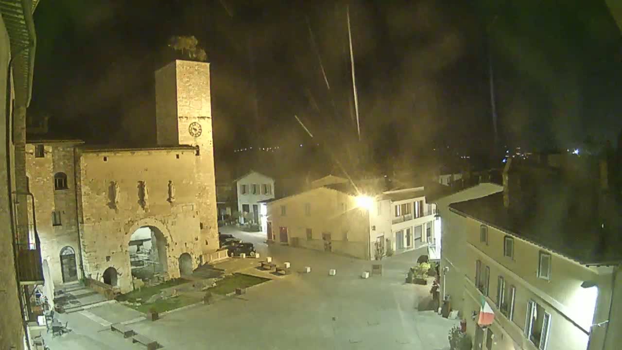 A nighttime view of a town square with old stone buildings, including a tall clock tower, under a partly cloudy sky with streaks of light possibly from fireworks.