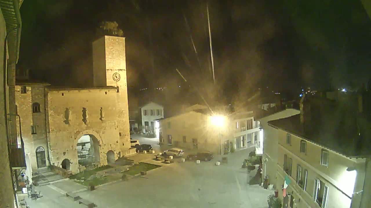 A nighttime view of a town square, featuring a tall clock tower, stone buildings, and a few parked cars under a hazy sky with streaks of light possibly from fireworks or shooting stars.