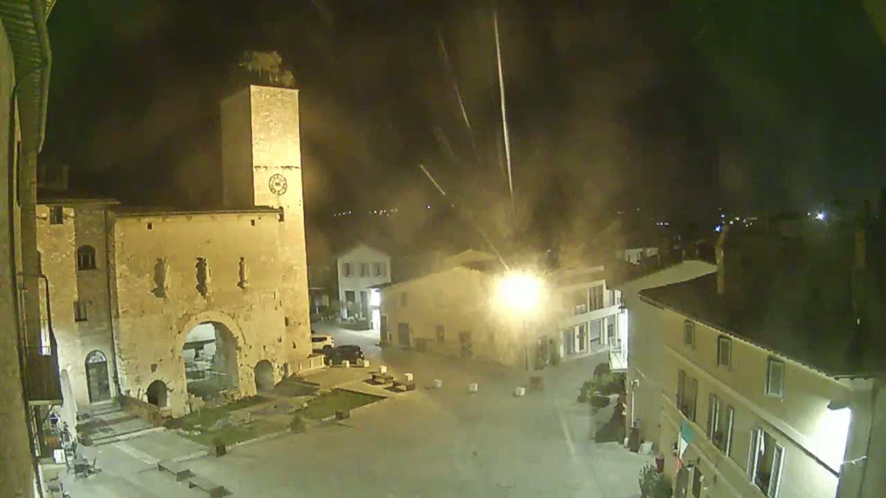 A nighttime view of a town square with ancient stone buildings, including a tall clock tower, under hazy conditions with faint streaks of light in the sky.