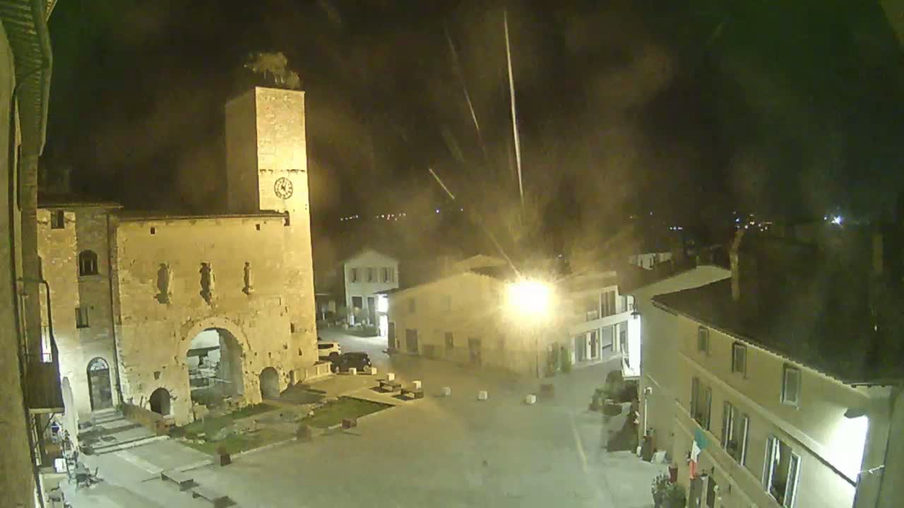 A nighttime view of a town square with stone buildings, including a tall clock tower, illuminated by streetlights under a hazy sky.