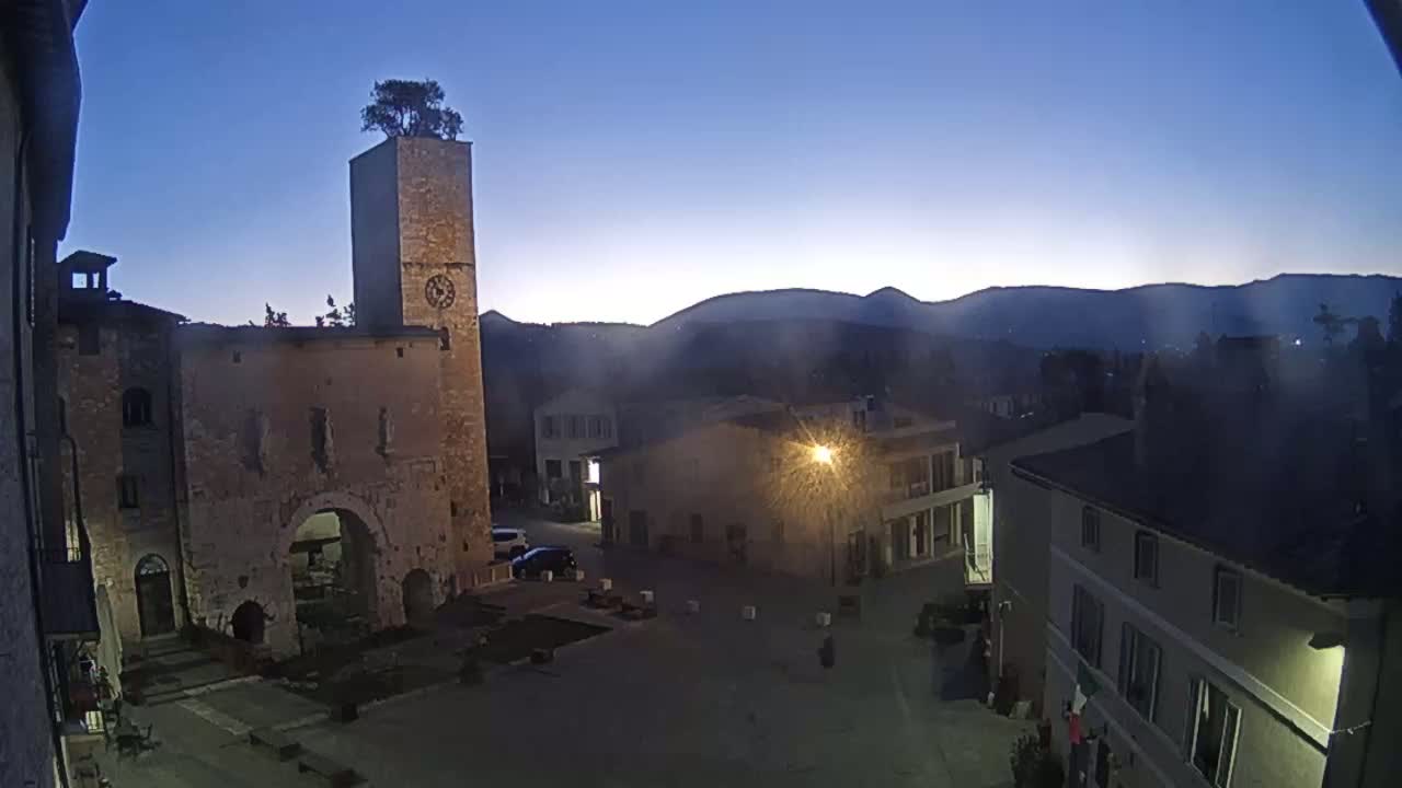 A town square at twilight, featuring a stone tower with a tree on top, is surrounded by old buildings against a backdrop of mountains.