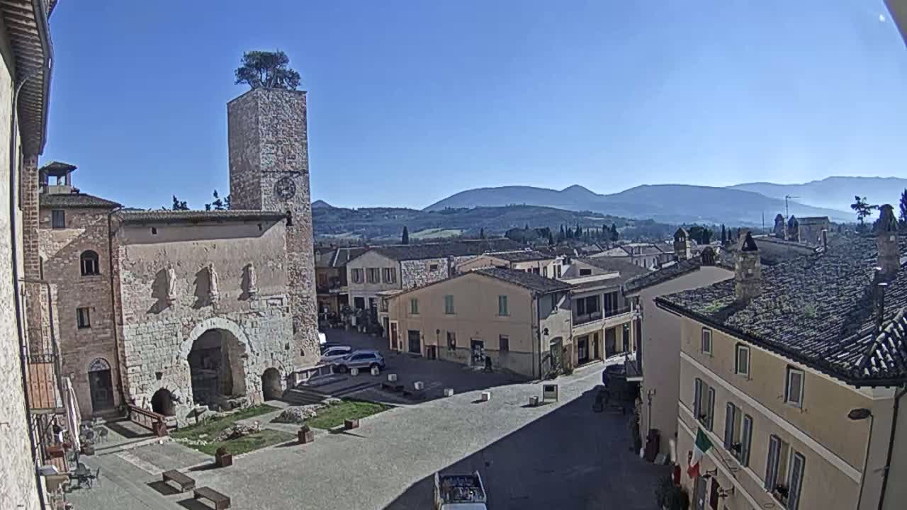A town square in a small Italian village on a clear, sunny day, is surrounded by old stone buildings, one with a clock tower topped with a tree, and overlooks rolling hills in the distance.