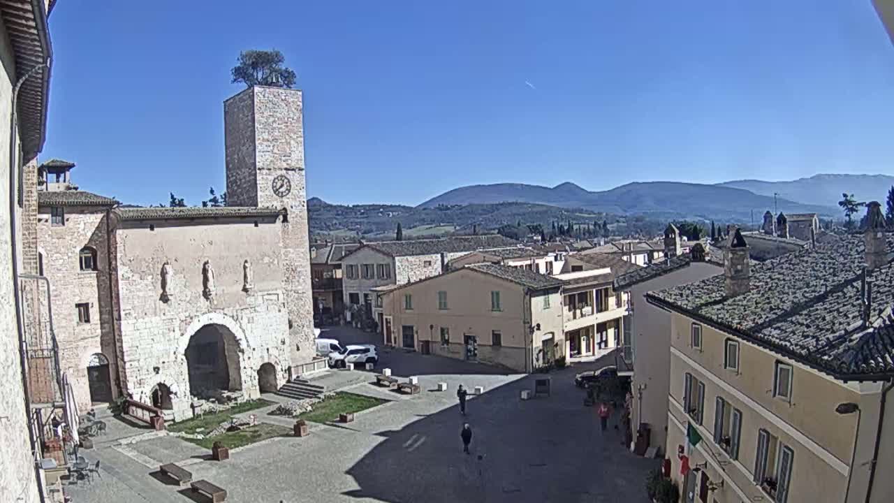 A town square with a clock tower and stone buildings, under a clear blue sky, overlooks rolling hills in the distance.
