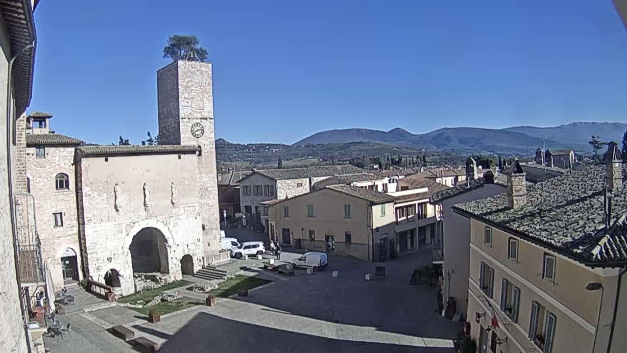 A town square in a small Italian town, under a clear blue sky, is surrounded by stone buildings, one featuring a tall tower with a tree on top, and overlooks distant mountains.