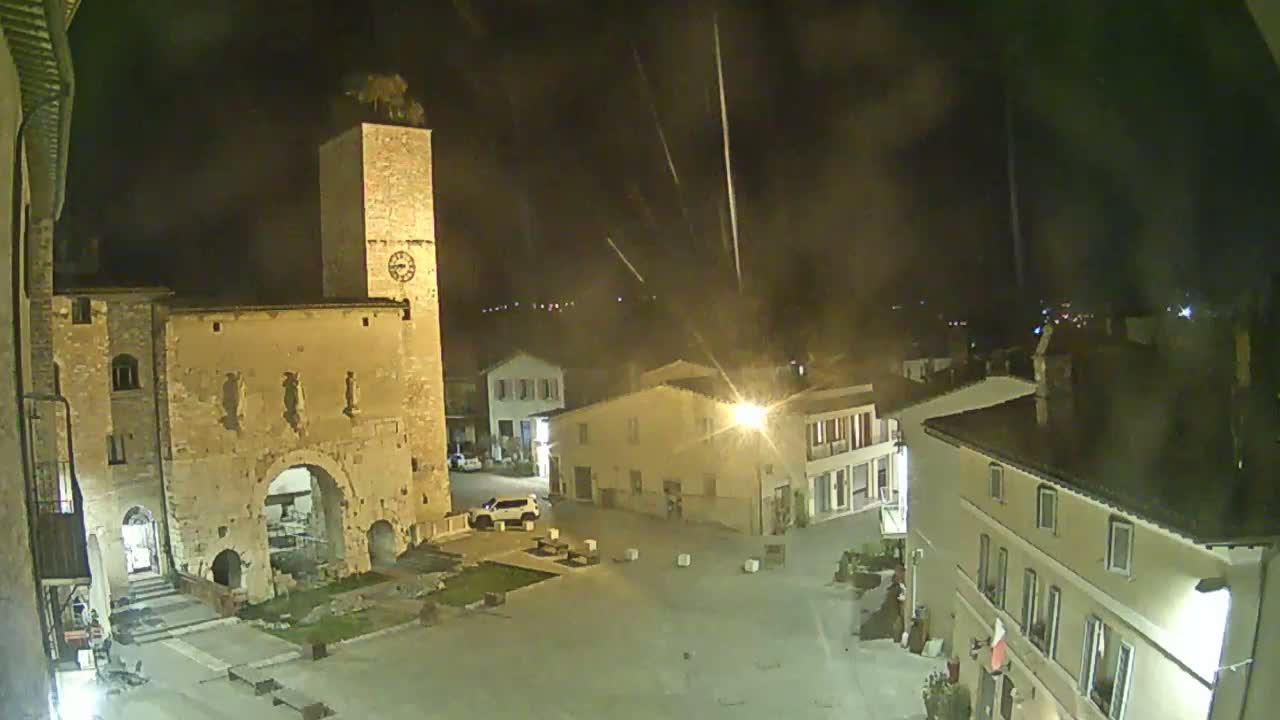 A nighttime view of a town square with a clock tower and several buildings illuminated under a dark, possibly hazy sky.