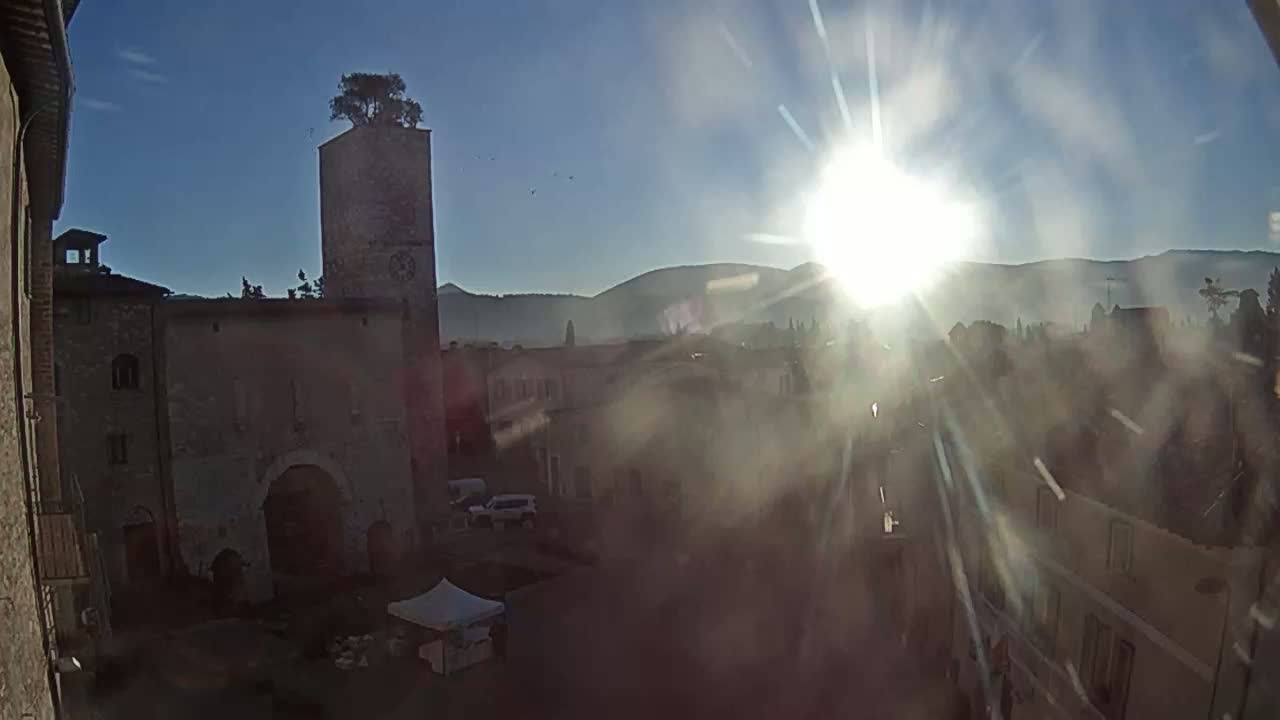 A sun-drenched town square is viewed from above, showing old stone buildings, a tall tower with a tree on top, and a bright sun causing lens flare.