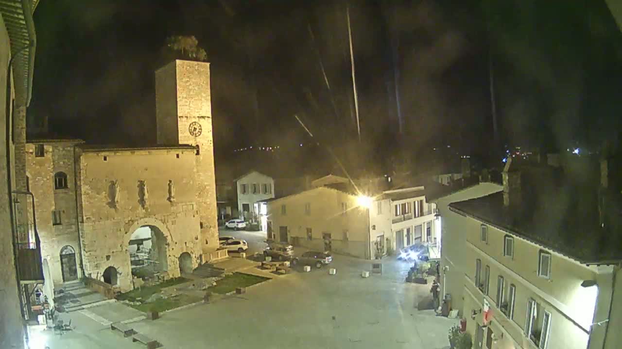A nighttime view of a town square with several buildings, including a tall stone tower, under a clear sky with light streaks possibly from fireworks.