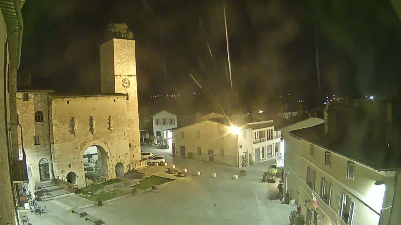 A nighttime view of a town square with a clock tower, stone buildings, and a few cars parked, under a slightly hazy sky with streaks of light possibly from fireworks.