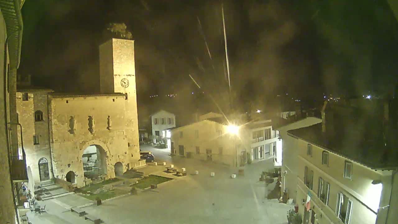 A nighttime, high-angle view of a town square with ancient stone buildings, a clock tower, and light streaks in the sky.