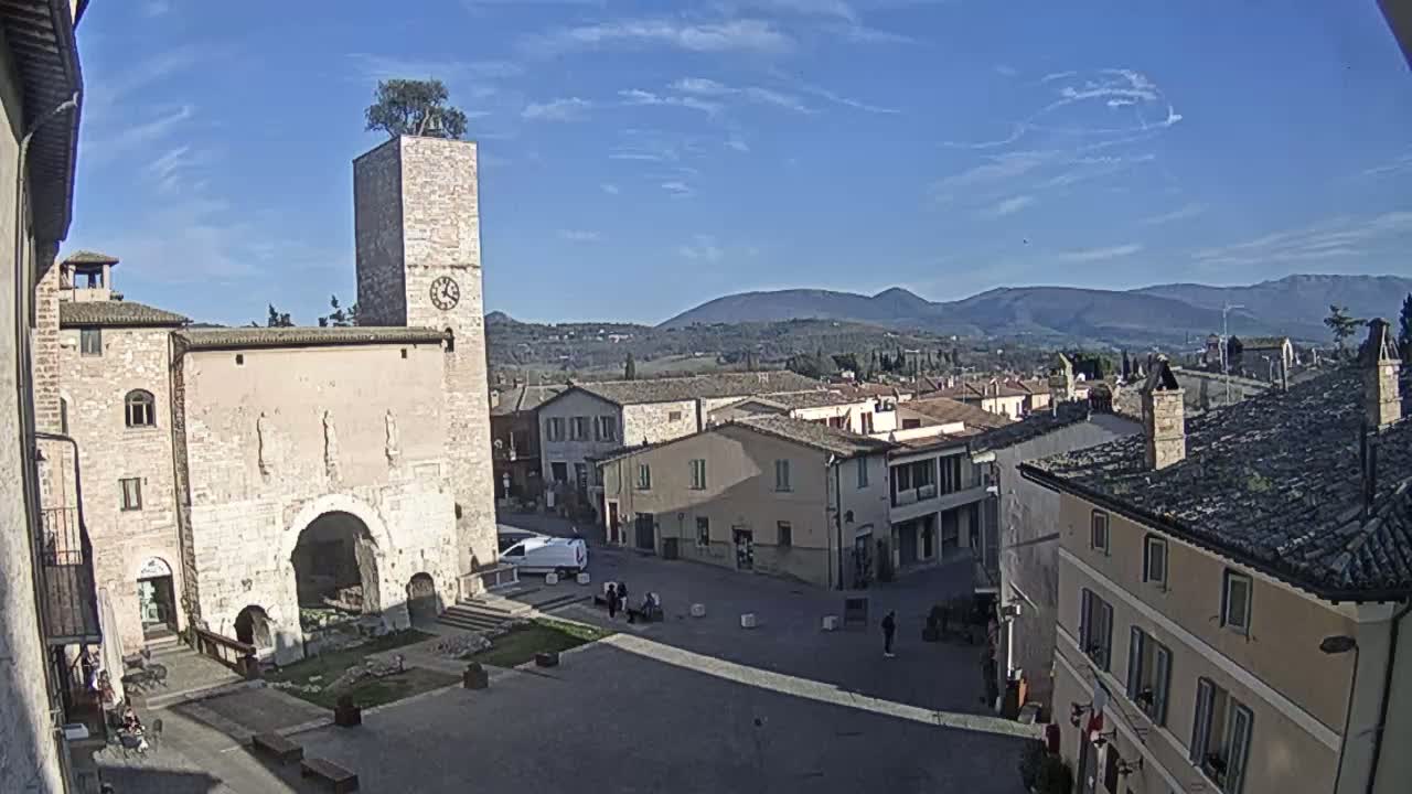 A town square with stone buildings, a clock tower topped with a tree, and distant mountains under a clear blue sky.