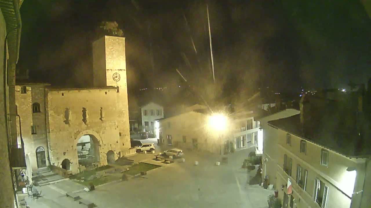 A nighttime view of a town square with an old stone building and clock tower, under a slightly hazy sky with streaks of what appears to be rain or falling debris.