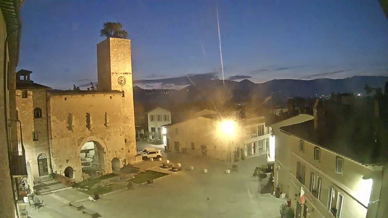 A town square at twilight, illuminated by streetlights, features a tall stone tower with a tree on top and several buildings under a clear, dark blue sky.
