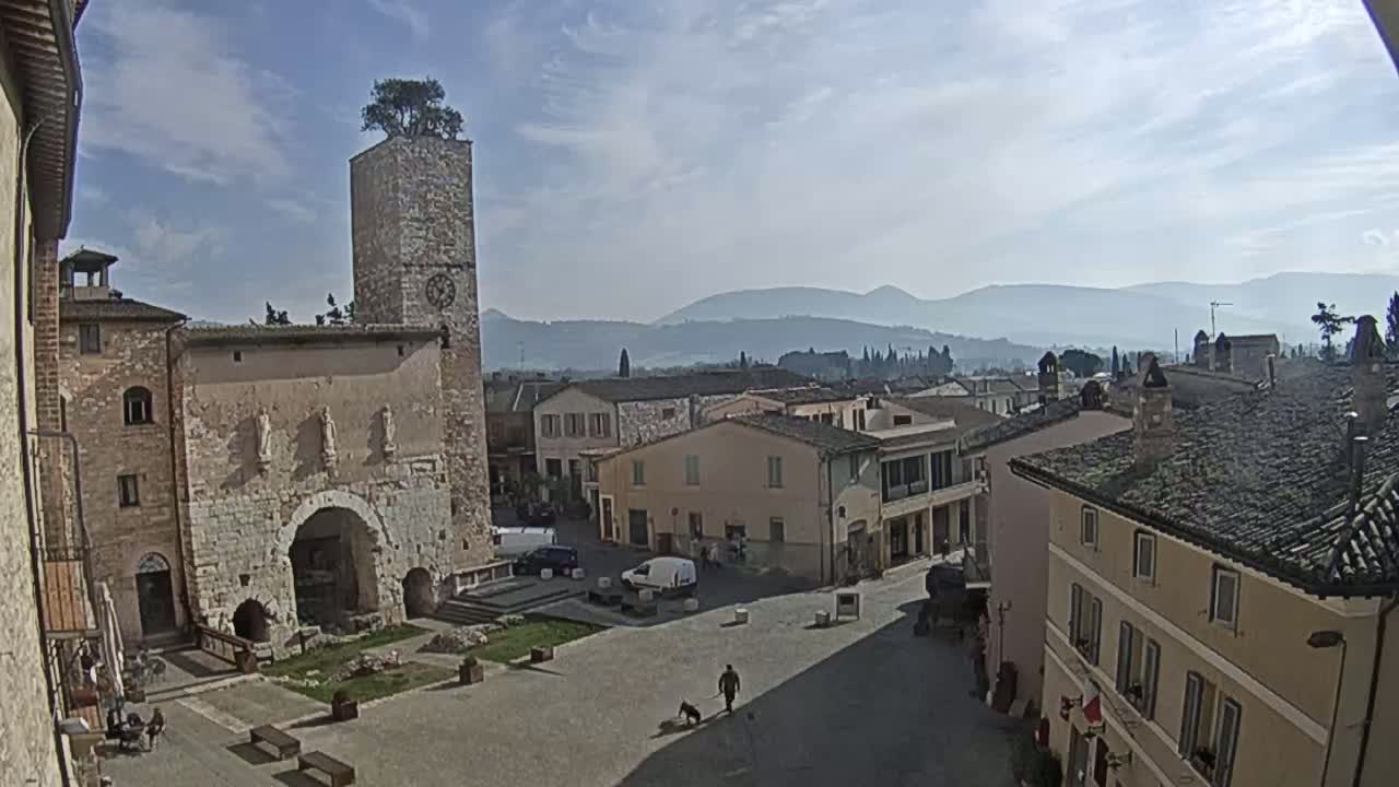 A mostly sunny view of a town square in a historic European town, with a stone tower, buildings, and distant mountains under a partly cloudy sky.