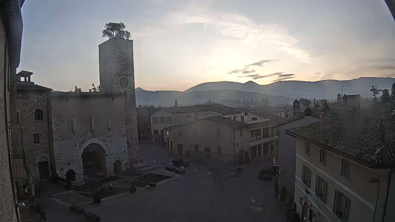 A town square with stone buildings, a tall tower with a tree on top, and distant mountains under a hazy sunrise.